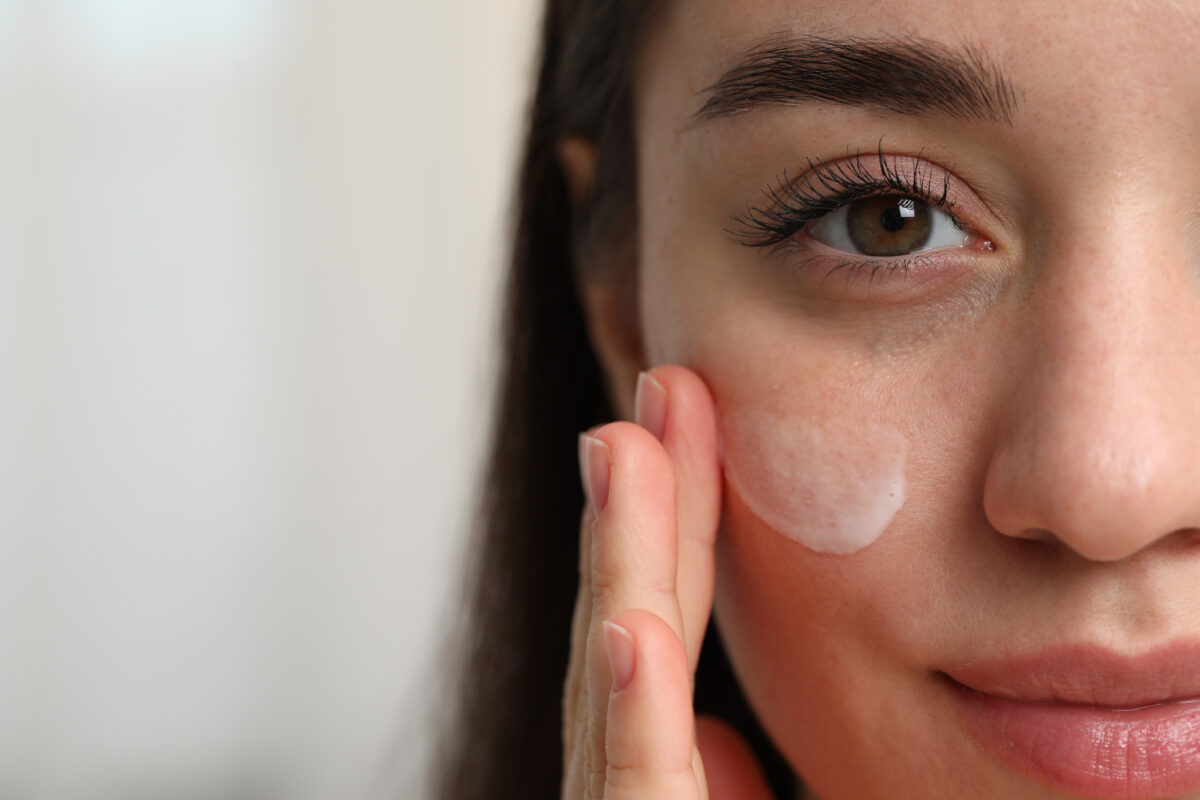 Young woman with dry skin applying cream onto her face on blurred background, closeup. Space for text